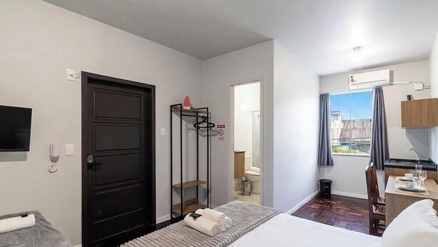 A hotel room with a bed, a desk, a chair, a wardrobe, a shower area, and a window with a view. - Studio in Velha w/ City View Ola0002 (Blumenau)