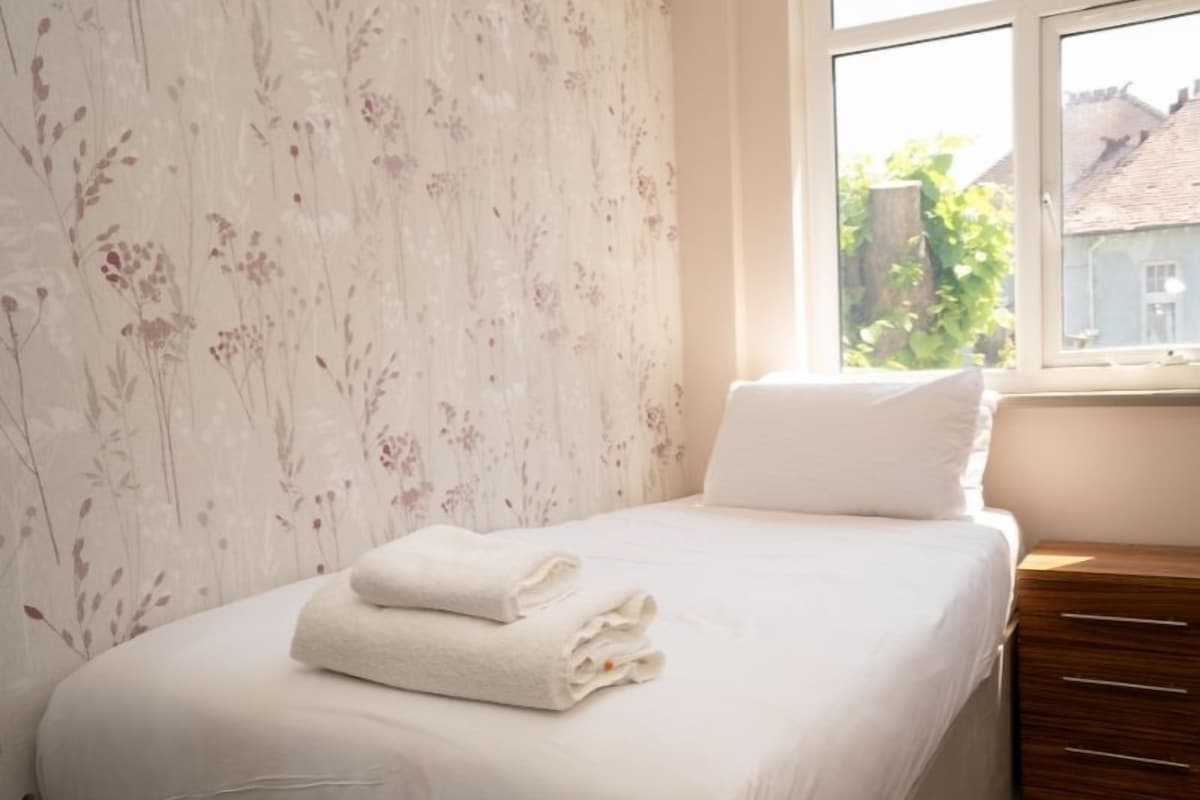 Una cama bien hecha con sábanas blancas y una toalla doblada, una mesita de noche de madera a la derecha, y una ventana con vistas a edificios y zonas verdes.