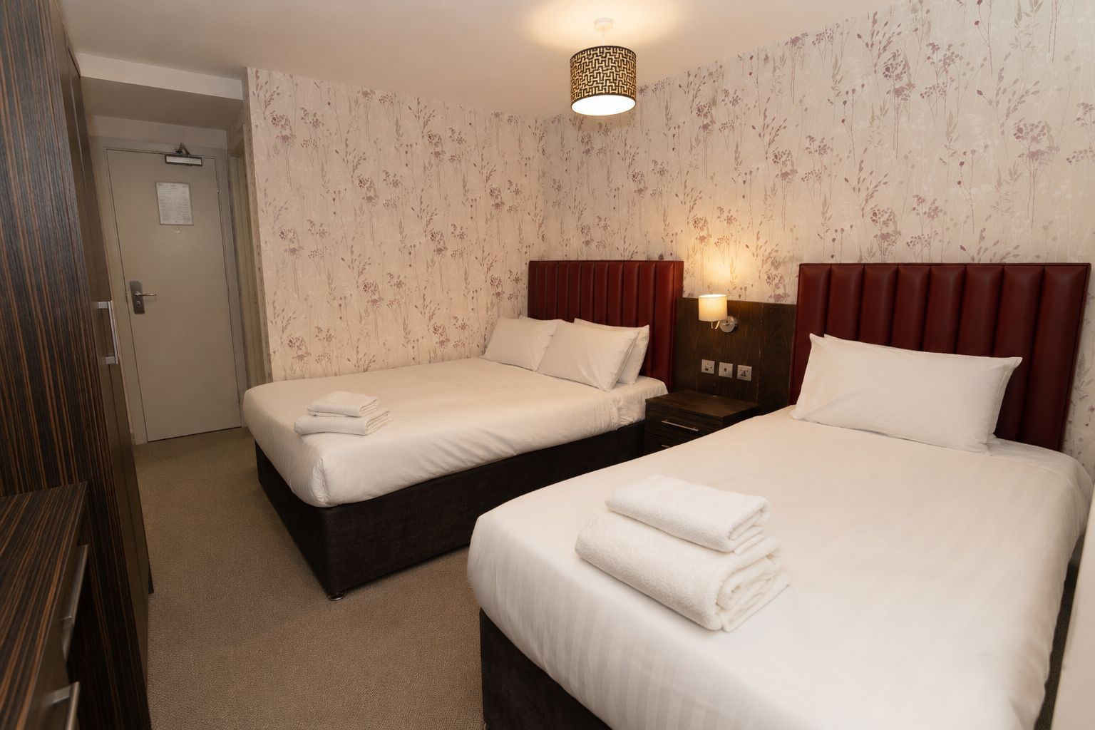 A hotel room with two beds, a door, and a wall with floral wallpaper.