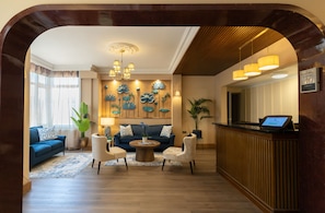A hotel lobby with a reception desk, seating area with sofas, a coffee table, and a decorative wall with floral artwork.