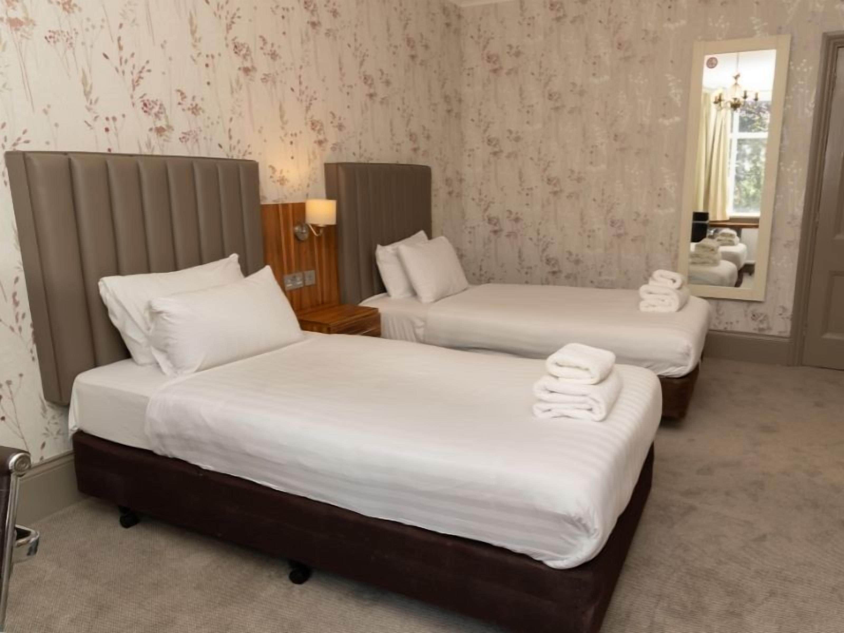 Quarto de hotel com duas camas, uma mesa de cabeceira, uma luminária e um espelho.
