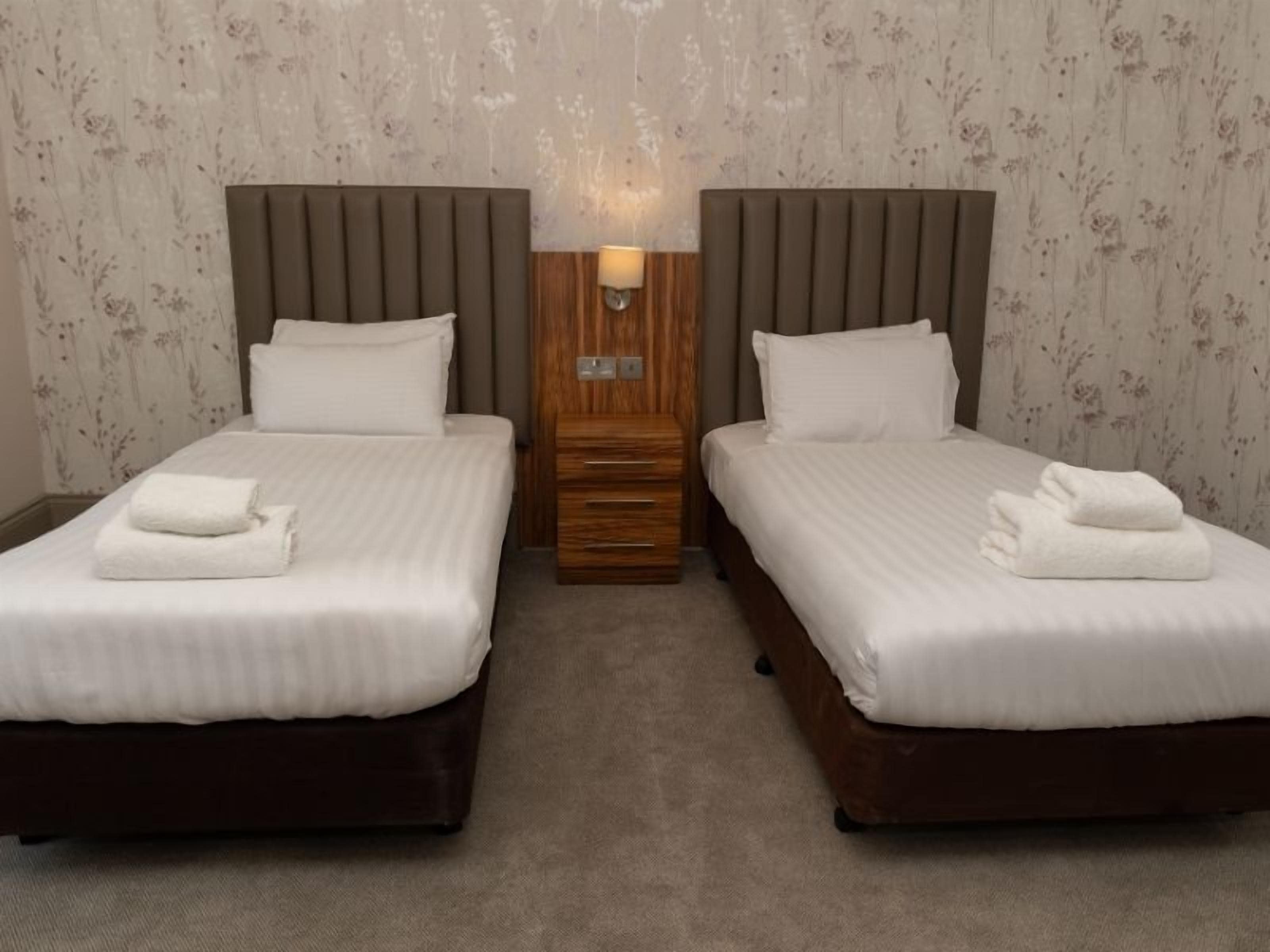 A hotel room with two beds, a wooden nightstand, and a wall-mounted lamp.