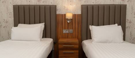 A hotel room with two beds, a wooden nightstand, a wall-mounted lamp, and a power outlet.