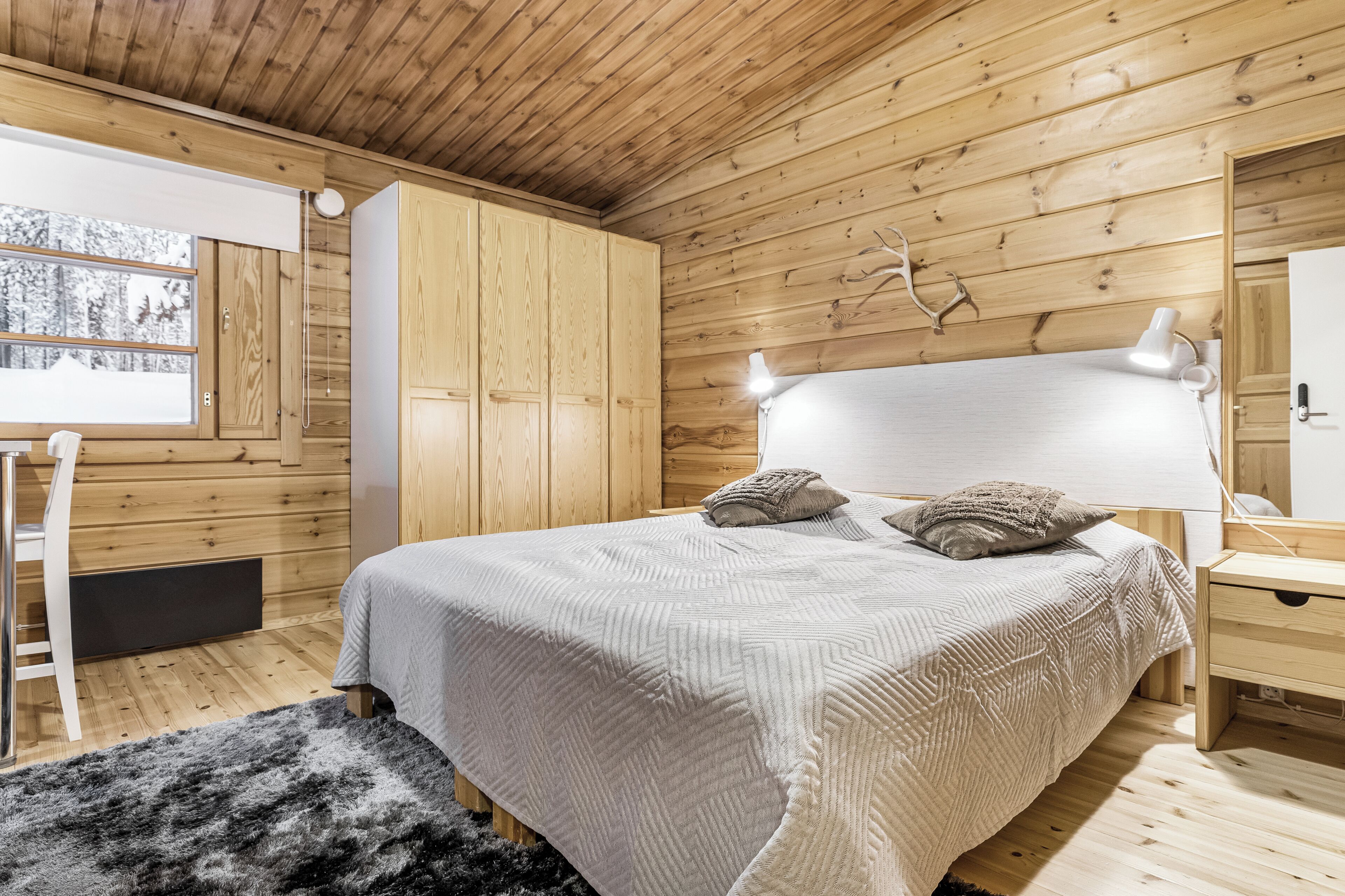 A bedroom with a wooden ceiling, a bed with a white headboard, a bedside table, a lamp, and a window with a view of trees.