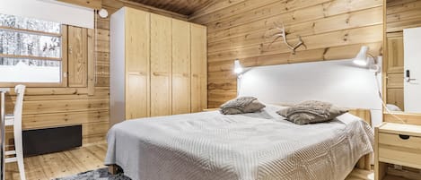 Ein Schlafzimmer mit Holzboden, einem Bett mit weißem Kopfteil, einem Nachttisch, einer Lampe und einem Fenster mit Blick auf Bäume.