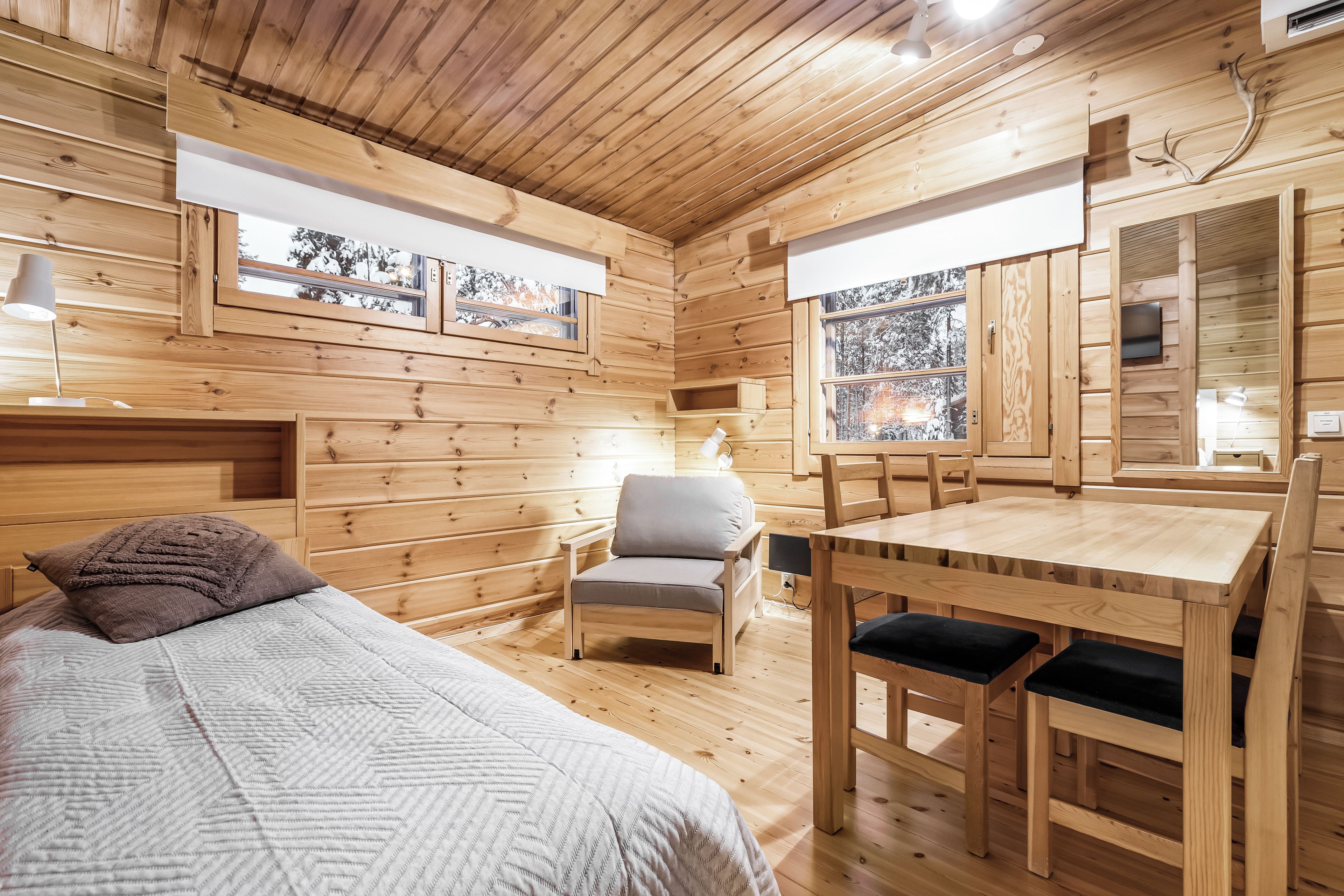 Ein Holzblock-Innenraum mit einem Bett, einem Tisch und Stühlen sowie großen Fenstern mit Blick auf Bäume.