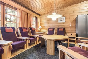 A wooden cabin interior with a dining table, chairs, a refrigerator, and a window with curtains.