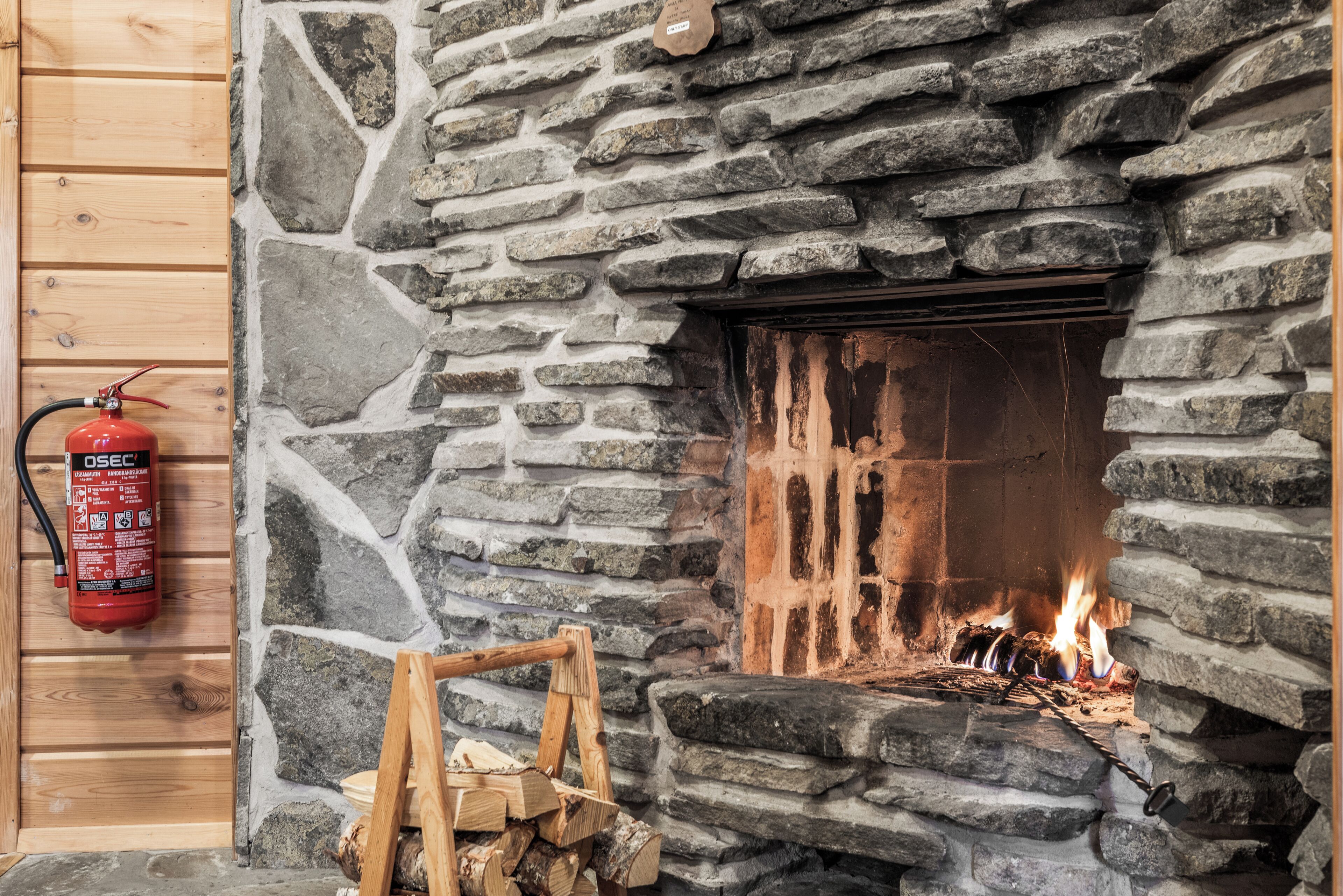 Une cheminée en pierre avec un feu qui crépite à l’intérieur, un extincteur fixé au mur sur la gauche, et une pile de bois de chauffage devant la cheminée.