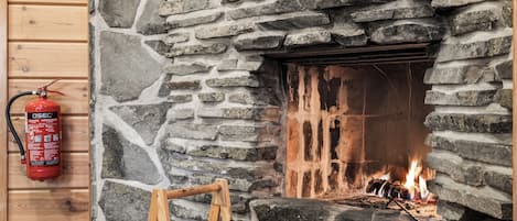 Ein Steinkamin mit brennendem Feuer, ein Feuerlöscher an der Wand links und ein Holzstapel vor dem Kamin.