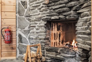 Una chimenea de piedra con fuego encendido en su interior, un extintor de incendios montado en la pared a la izquierda y una pila de leña frente a la chimenea.