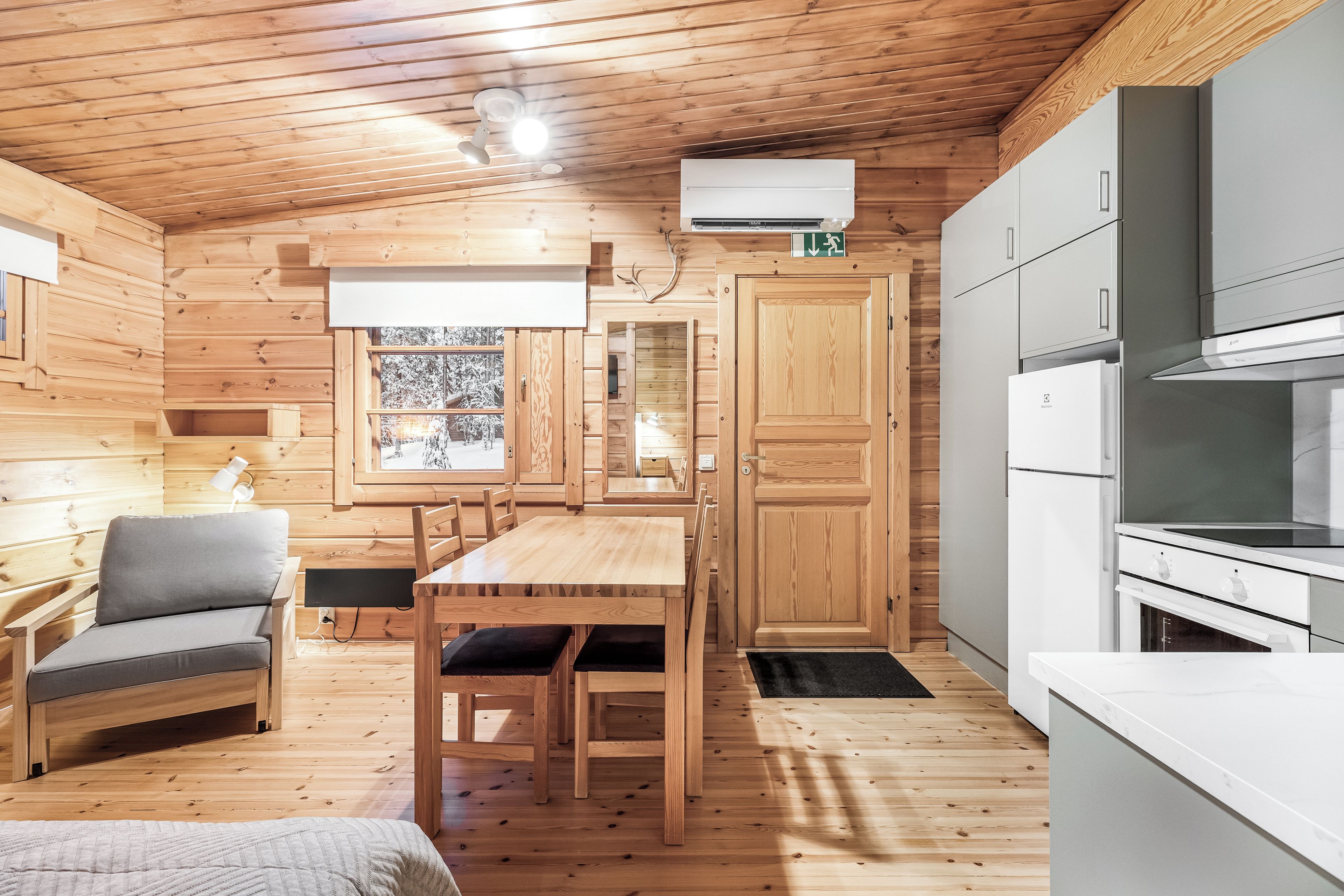 Un intérieur de chalet en bois, de taille réduite, comprenant un coin repas, une kitchenette et une fenêtre donnant sur des arbres couverts de neige.