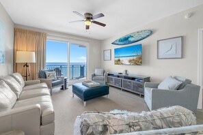 A spacious living area with a balcony overlooking the ocean, a ceiling fan, and a television. - Sterling Breeze 507- Dancing Waves (Panama City Beach)