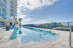 A swimming pool with a clear blue sky, a palm tree, and a building with balconies. - Sterling Breeze 507- Dancing Waves (Panama City Beach)
