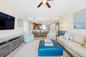 A living room with a TV, a blue ottoman, a beige sofa, and a ceiling fan. - Sterling Breeze 507- Dancing Waves (Panama City Beach)