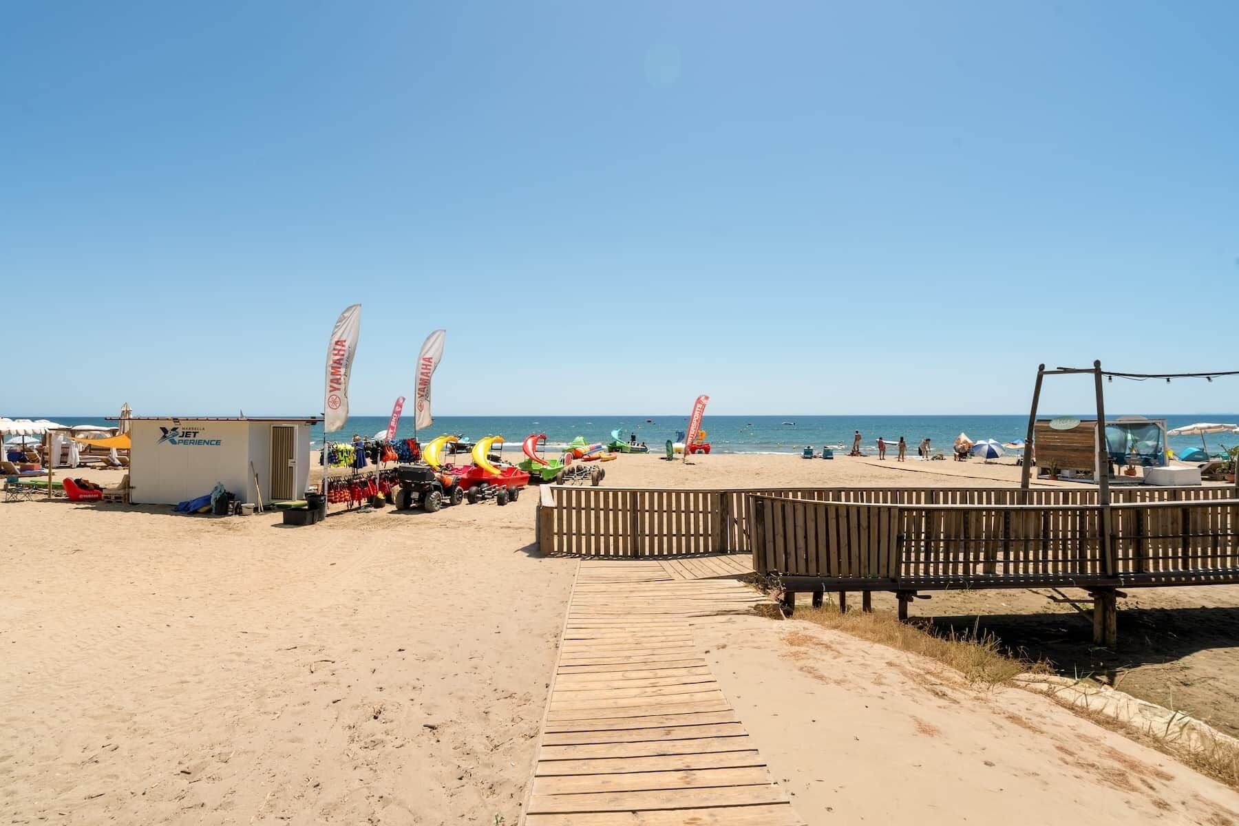 A beach with a wooden boardwalk leading to a sandy area, a small building, and flags.