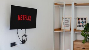 Televisor montado en la pared con el logo de Netflix, una estantería con libros y un juego de mesa, y un enchufe.