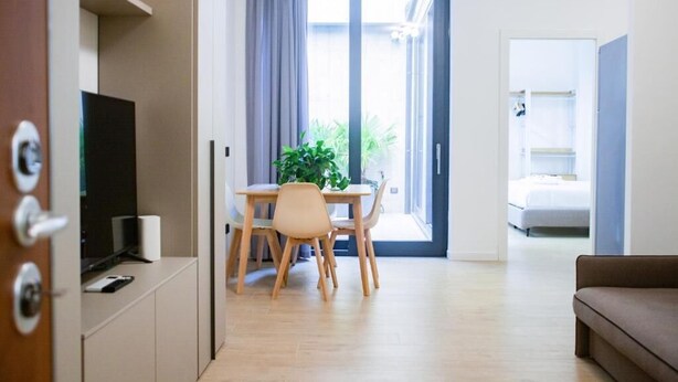 Una sala moderna con un sofá, un comedor con mesa y sillas, un televisor y un ventanal corredizo que da a un balcón.