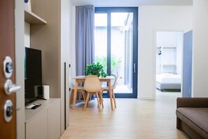 Una sala moderna con un sofá, un comedor con mesa y sillas, un televisor y un ventanal corredizo que da a un balcón.
