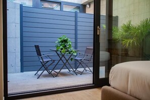 Ein Zimmer mit einer Schiebetür zum Balkon, der mit einem Tisch, Stühlen und einem Blumentopf möbliert ist. Ein blaues Geländer begrenzt den Außenbereich.