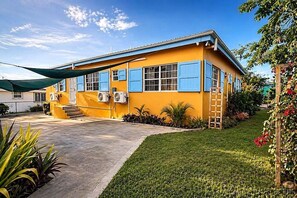 A yellow building with blue shutters, a concrete driveway, and a small garden with plants and a ladder. - 3-bedroom house- Close Prox to Airport, Beaches and Amenities full AC (Frederiksted)