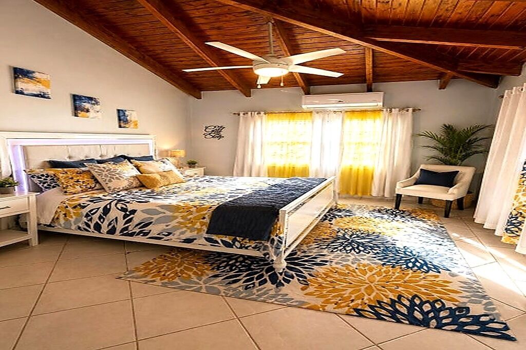 Ein Schlafzimmer mit einem Bett, einem Nachttisch, einem Sessel, einem Deckenventilator und einem Fenster mit Vorhängen.