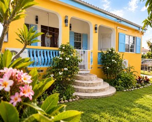 A yellow building with blue shutters, a staircase leading to the entrance, and a well-maintained garden with various plants and flowers.