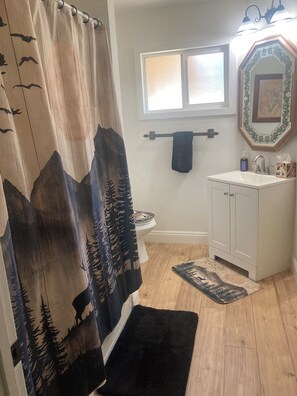 Un baño con cortina de ducha decorada con un diseño de montaña y bosque, un inodoro, un lavabo con dispensador de jabón, un perchero y una ventana sobre el lavabo.