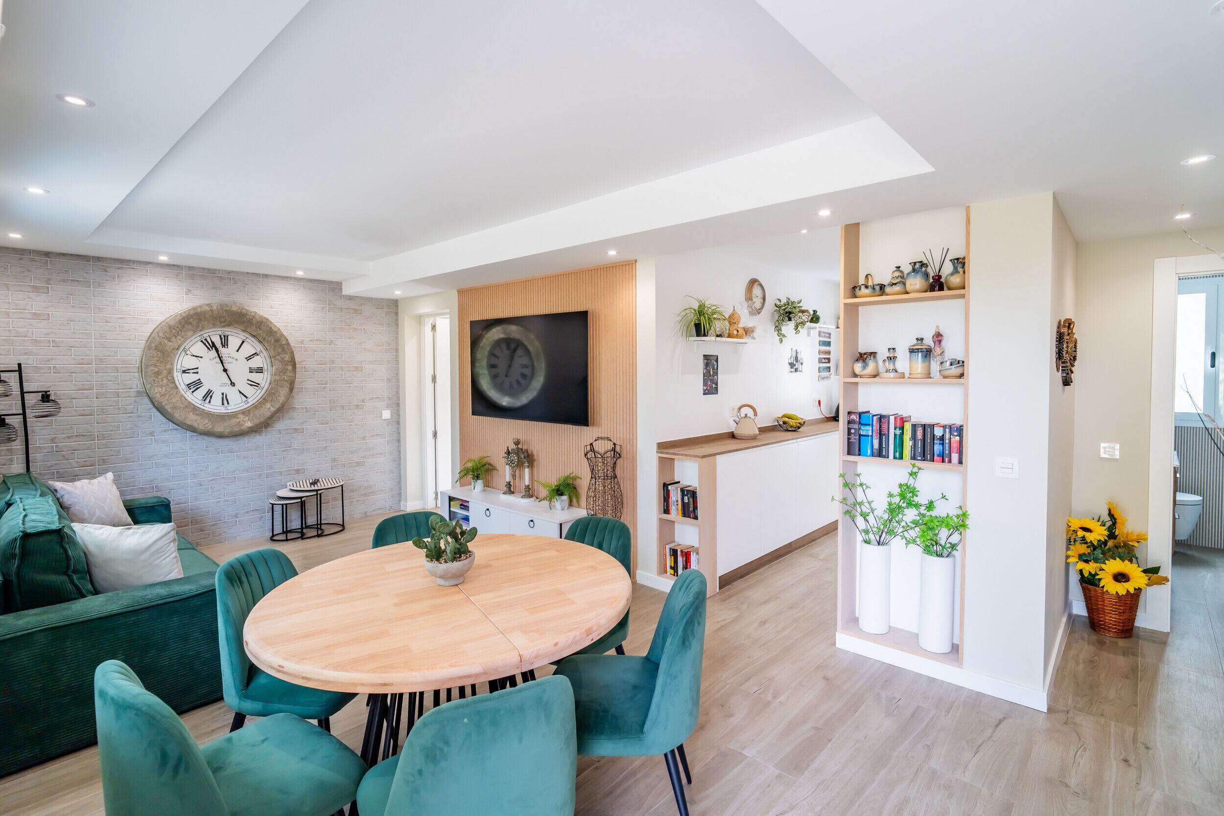 Una sala moderna con una mesa redonda de madera y sillas color turquesa, una pared de ladrillo con un reloj grande, y una zona de cocina con estantes y plantas.