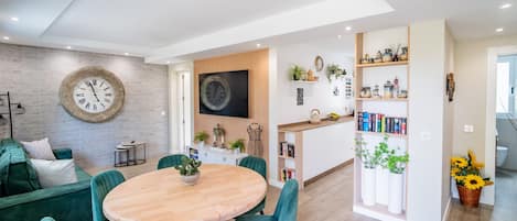 Una sala moderna con una mesa redonda de madera y sillas color turquesa, una pared de ladrillo con un reloj grande, y una zona de cocina con estantes y plantas.