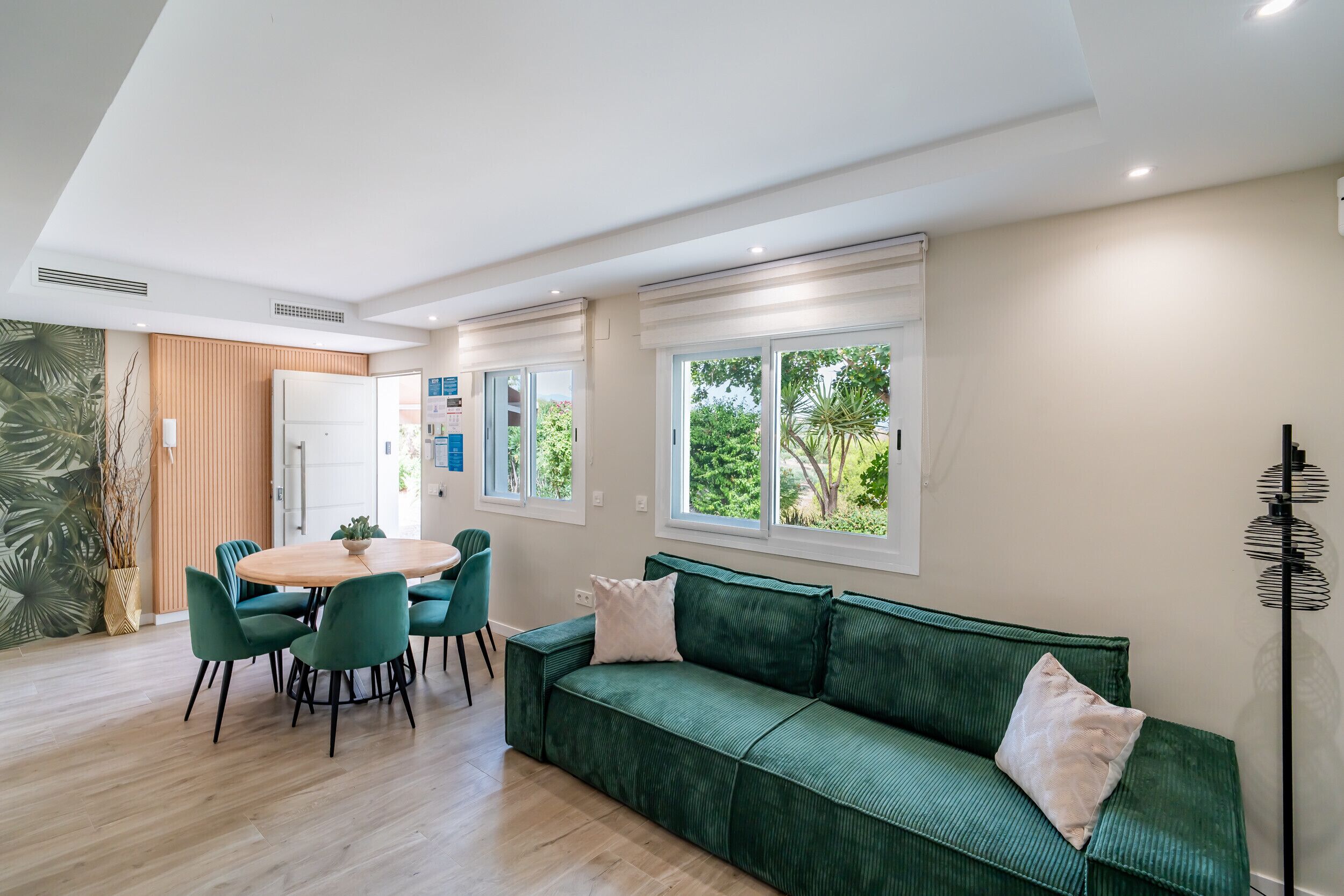 Ein modernes Wohnzimmer mit einem grünen Sofa, einem runden Esstisch mit Stühlen und großen Fenstern mit Blick auf Grünflächen.