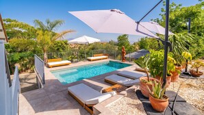 Piscina com espreguiçadeiras e guarda-sóis, rodeada por vegetação e um céu claro.