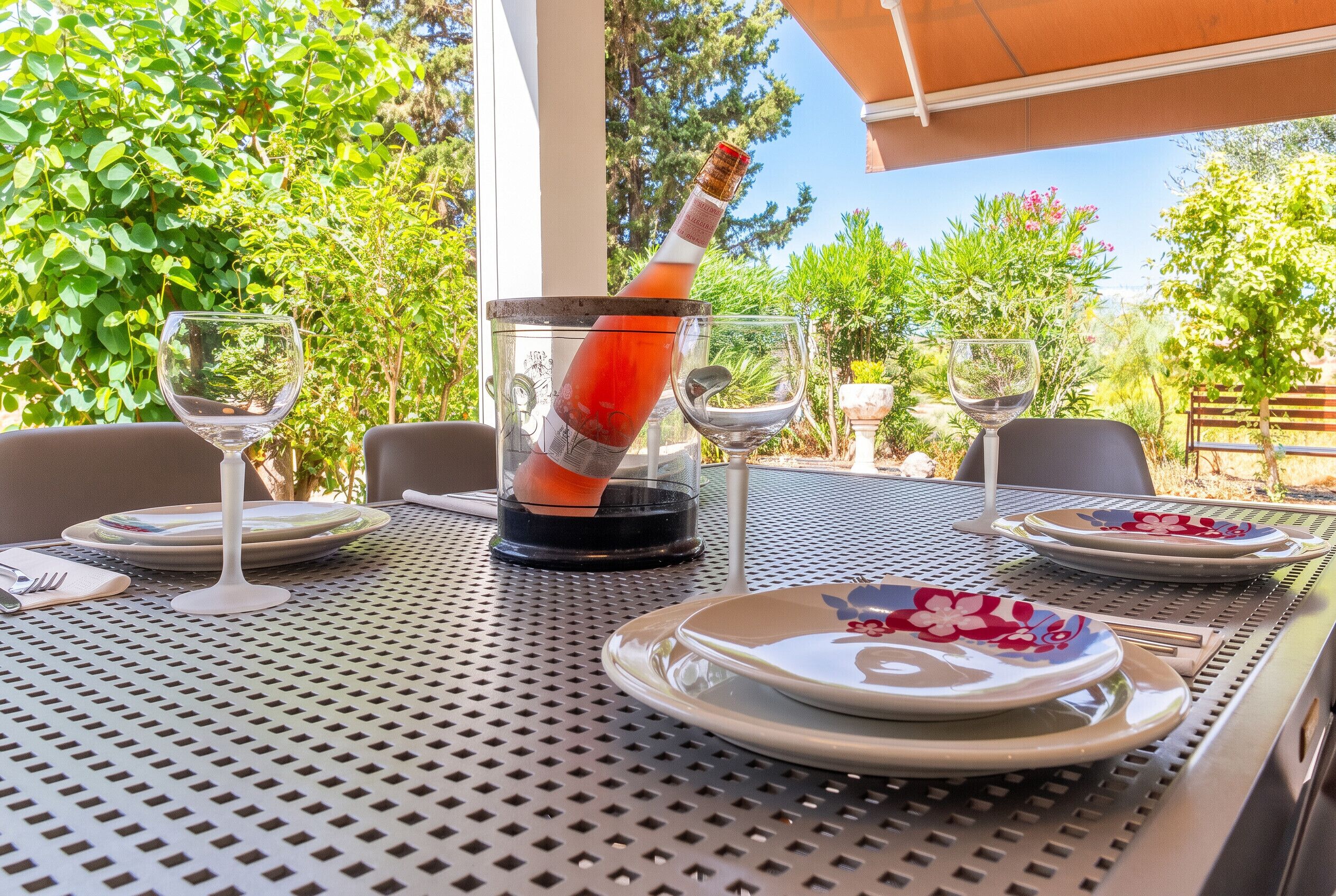 Un área de comedor al aire libre con una mesa preparada para dos personas, con sillas de metal, platos y copas de vino.