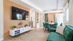 Uma sala de estar moderna com televisão de tela plana na parede, um móvel para eletrônicos de madeira, um espaço de jantar com mesa e cadeiras, e um sofá verde.