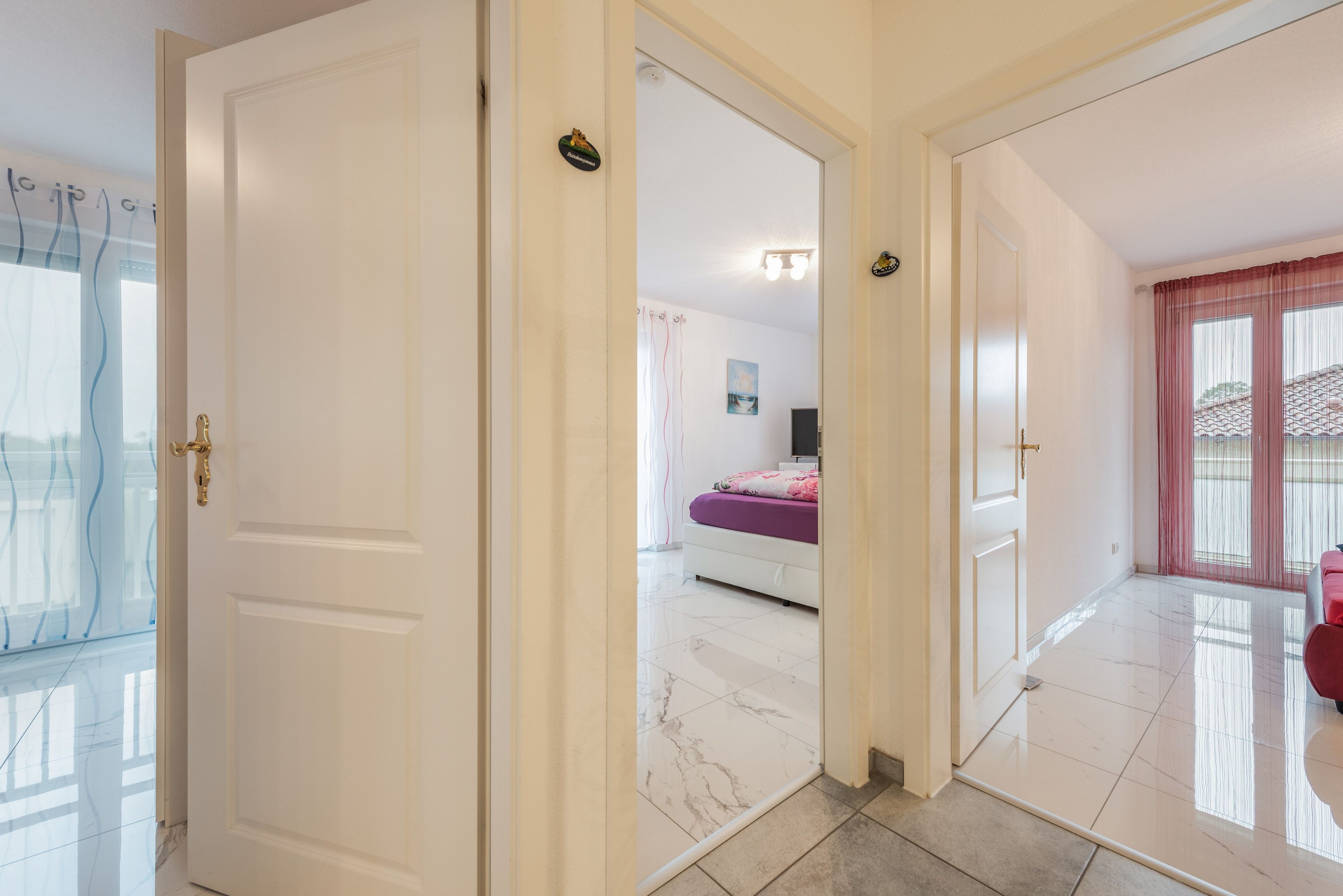 Ein Flur, der zu einem Schlafzimmer mit Bett, einem fensterbelaubten Fenster und einer Tür mit Griff führt.