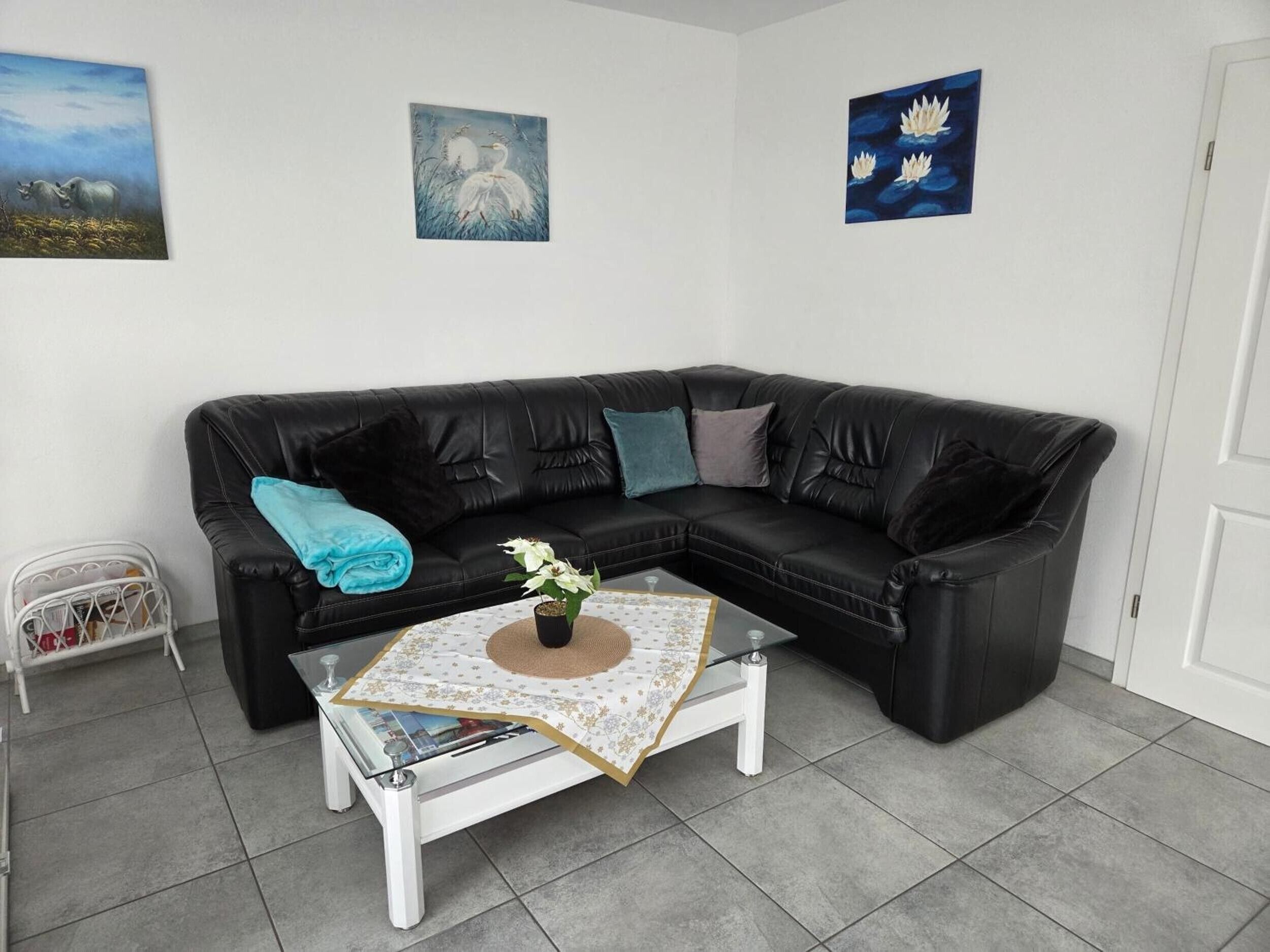 Ein Wohnzimmer mit einem schwarzen Ledersofa, einem Glastisch mit floralem Untersetzer und zwei Wandbildern.