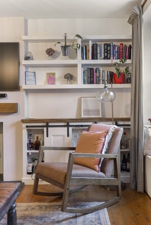 Ein gemütliches Wohnzimmer mit einem Holzschaukelstuhl, einem Bücherregal voller Bücher, einem Flachbildfernseher und einer kleinen Pflanze.