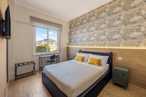 Ein Schlafzimmer mit Bett, Schreibtisch, Stuhl, Fernseher und einem Fenster mit Blick auf Gebäude.