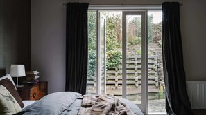 Ein Schlafzimmer mit einem Bett, einem Nachttisch, einer Lampe und Blick durch eine Schiebetür auf einen Außenbereich mit Holzzaun und Grünflächen.