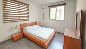 A bedroom with a wooden bed, a nightstand, a lamp, a window with blinds, and a ceiling light. - Modern 1BR Apartment in Strovolos (Strovolos)