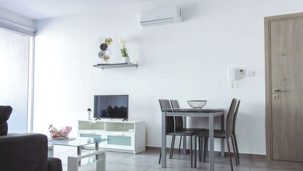 A modern living room with a flat-screen TV, a dining table with chairs, a white cabinet, and a wooden door. - Modern 1BR Apartment in Strovolos (Strovolos)
