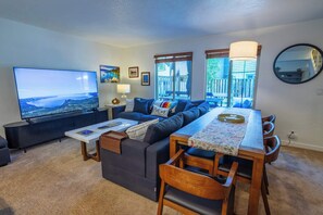 Ein modernes Wohnzimmer mit einem großen Flachbildfernseher, einer Couch, einem Esstisch mit Stühlen und Blick ins Freie durch große Fenster.