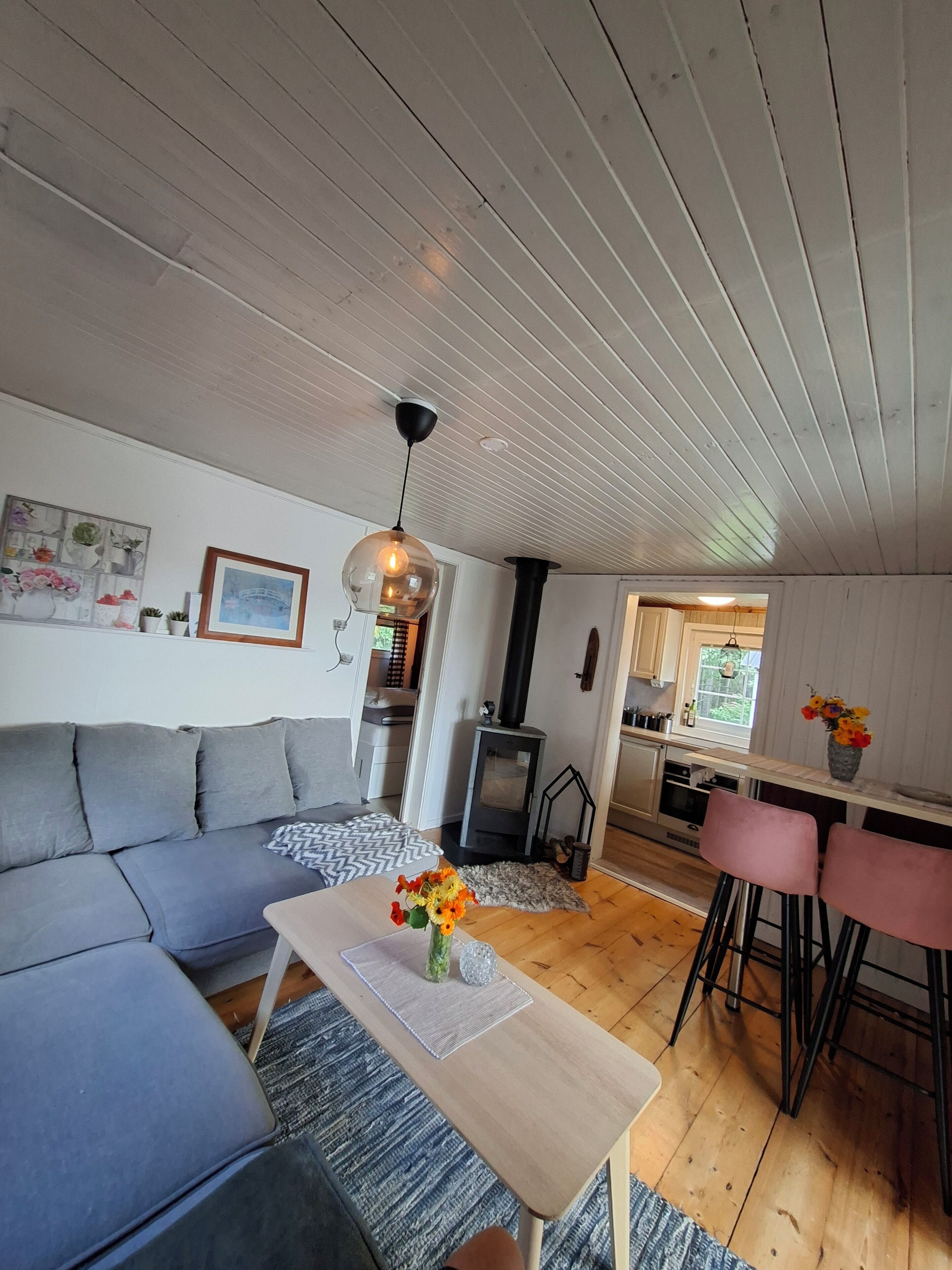 A modern living room with a grey sofa, a wooden coffee table with a vase of flowers, a hanging light fixture, and a kitchen area visible through an open doorway.