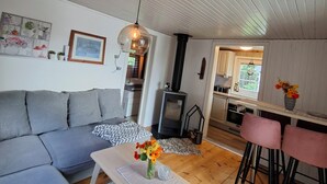 Una sala de estar moderna con un sofá gris, una mesa de centro de madera con un florero, una lámpara colgante y una cocina visible a través de un vano abierto.