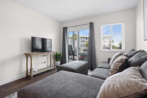 Un salon moderne avec un téléviseur à écran plat posé sur une console en bois, des canapés gris, une petite table d’appoint avec une plante, et une vue sur un quartier résidentiel grâce à de grandes fenêtres.
