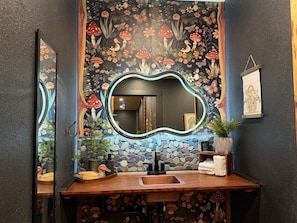 A bathroom with a stone mosaic wall, a wooden vanity, a mirror with a wavy frame, and a potted plant. - Fern & Feather No 1 at Cedar Creek Crossing (Mena)