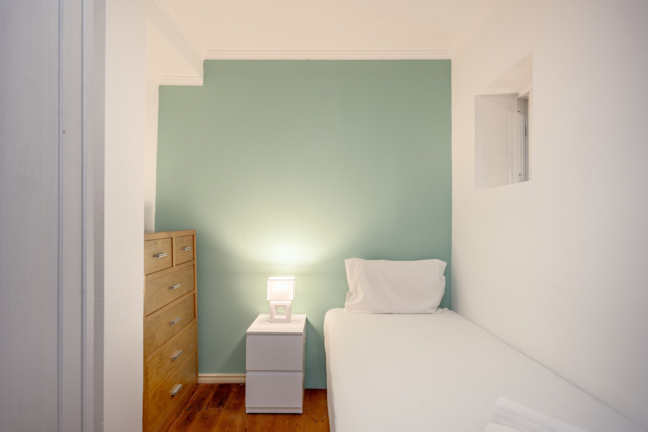 A small, narrow room with a single bed, a nightstand with a lamp, and a wooden dresser.