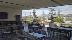 Ein Außenbereich mit einer Terrasse, Tischen und Stühlen, einem an der Wand befestigten Flachbildfernseher und Blick auf Stadt und Berge im Hintergrund.