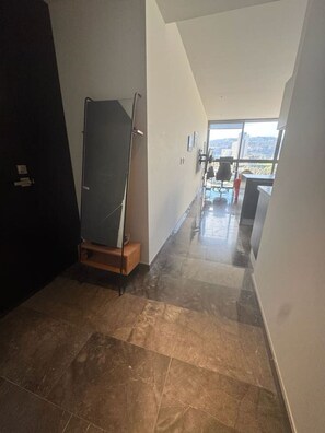 Un couloir avec une grande fenêtre, une porte et un miroir posé sur un piédestal.