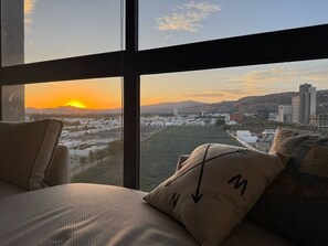 Ein Zimmer mit großem Fenster und Blick auf eine Stadtlandschaft bei Sonnenuntergang.