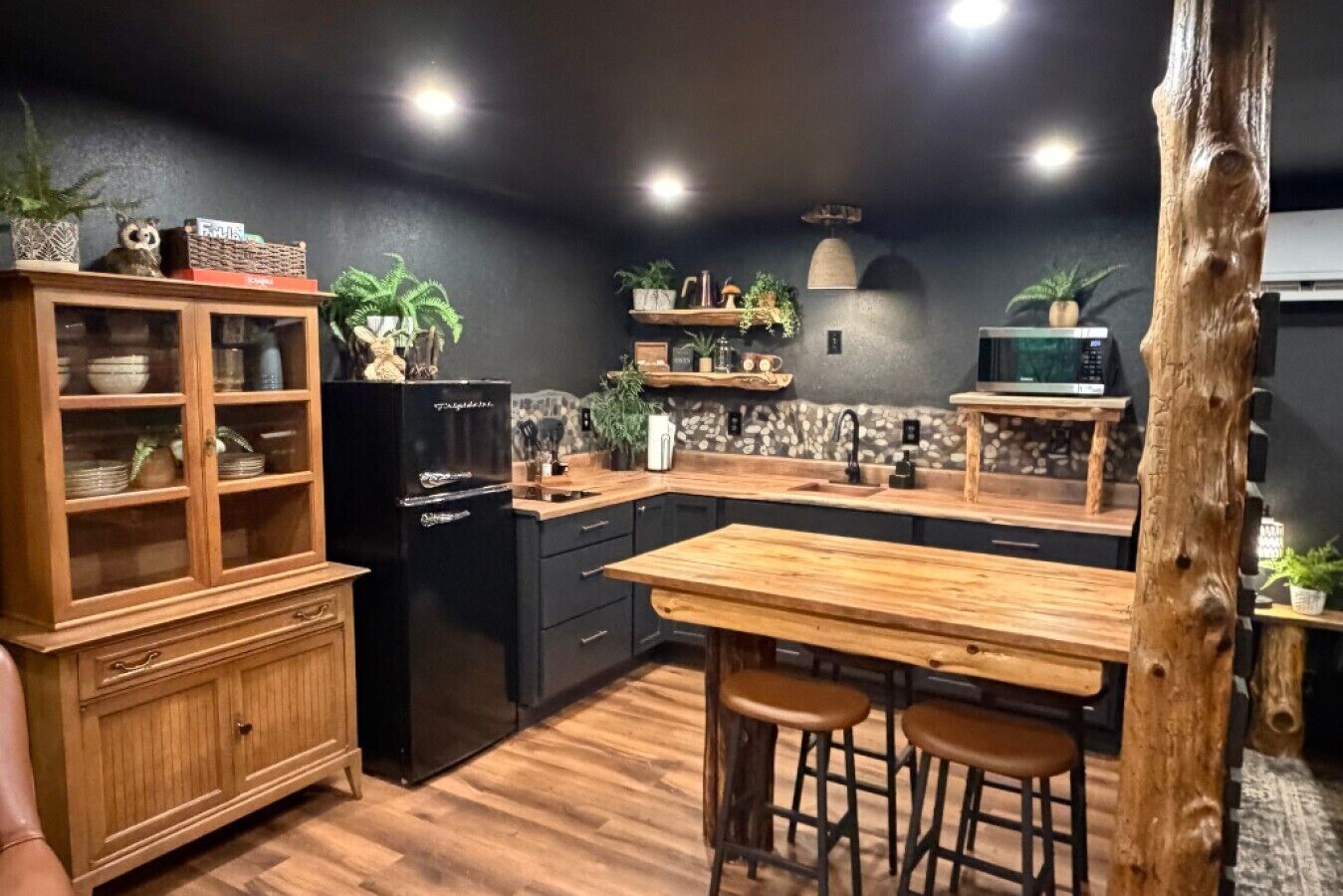 Une cuisine avec des meubles de rangement en bois, un réfrigérateur noir, une table à manger en bois avec des tabourets et diverses plantes.